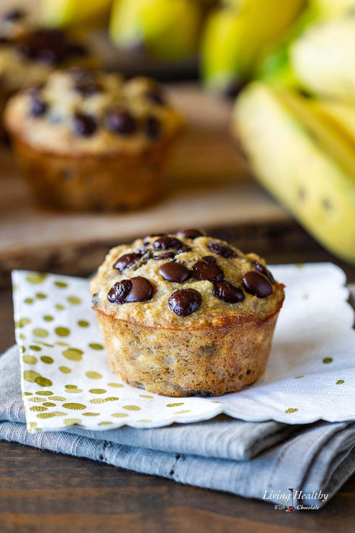 close up of coconut banana muffin topped with chocolate chips on stack of multicolored napkins with banana and more muffins in background