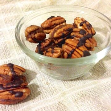 small glass dish with chocolate drizzled caramel filled pecans and a few loose pecans on the left side of the dish
