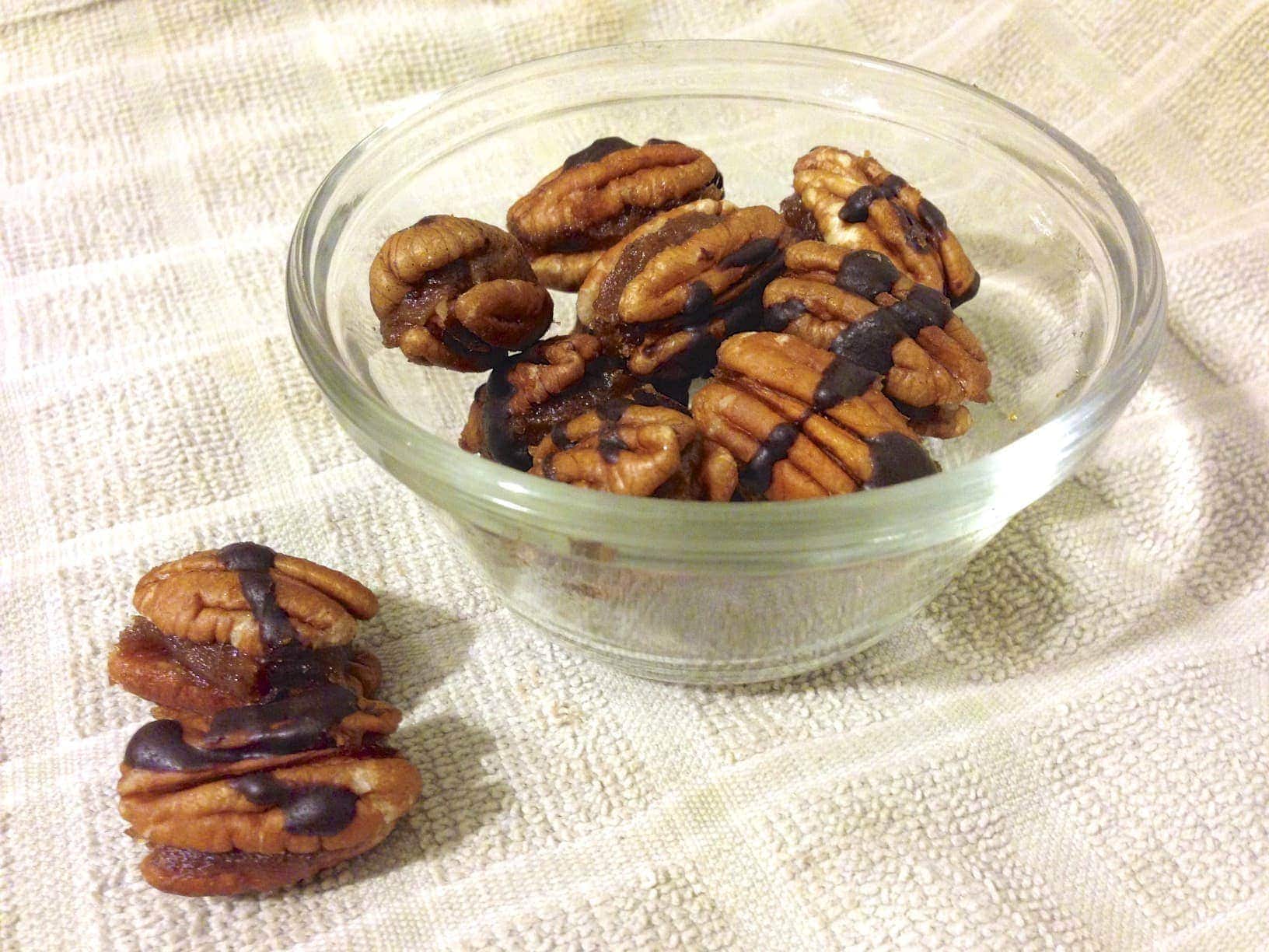 small glass dish with chocolate drizzled caramel filled pecans and a few loose pecans on the left side of the dish