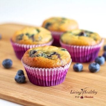 wooden cutting board topped with four coconut flour blueberry muffins in colorful muffin cups with loose blueberries around