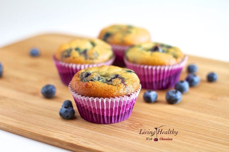 wooden table topped with four coconut flour blueberry muffins in colorful muffin cups with loose blueberries around