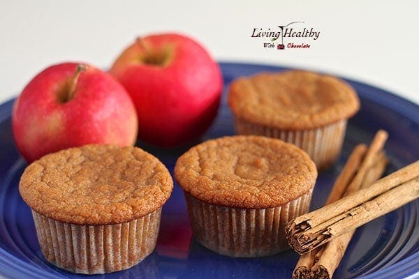 three apple cinnamon muffins on a blue plate with two red apples in background and sticks of cinnamon in foreground 