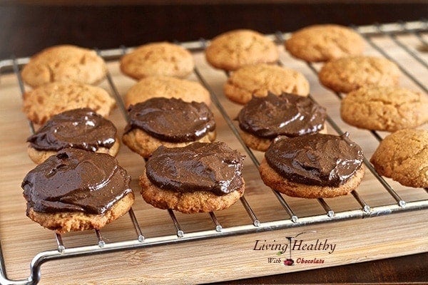 cooling rack with several rows of almond cookies topped with chocolate avocado fudge frosting 