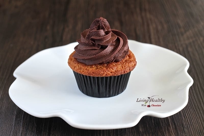 close up of a vanilla cupcake with chocolate banana frosting on a white plate sitting on a dark wooden table