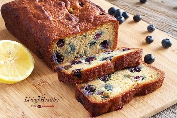 close up of paleo blueberry bread loaf with lemon glaze on cutting board with loose blueberries in background 