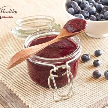 glass jar of sugar free blueberry butter with wooden spoon on top and bowl of blueberries in background