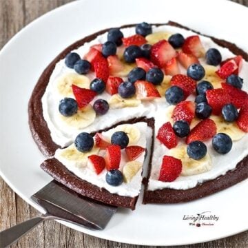 large serving plate with raw chocolate brownie fruit pizza dessert topped with cream and berries with one slice on spatula