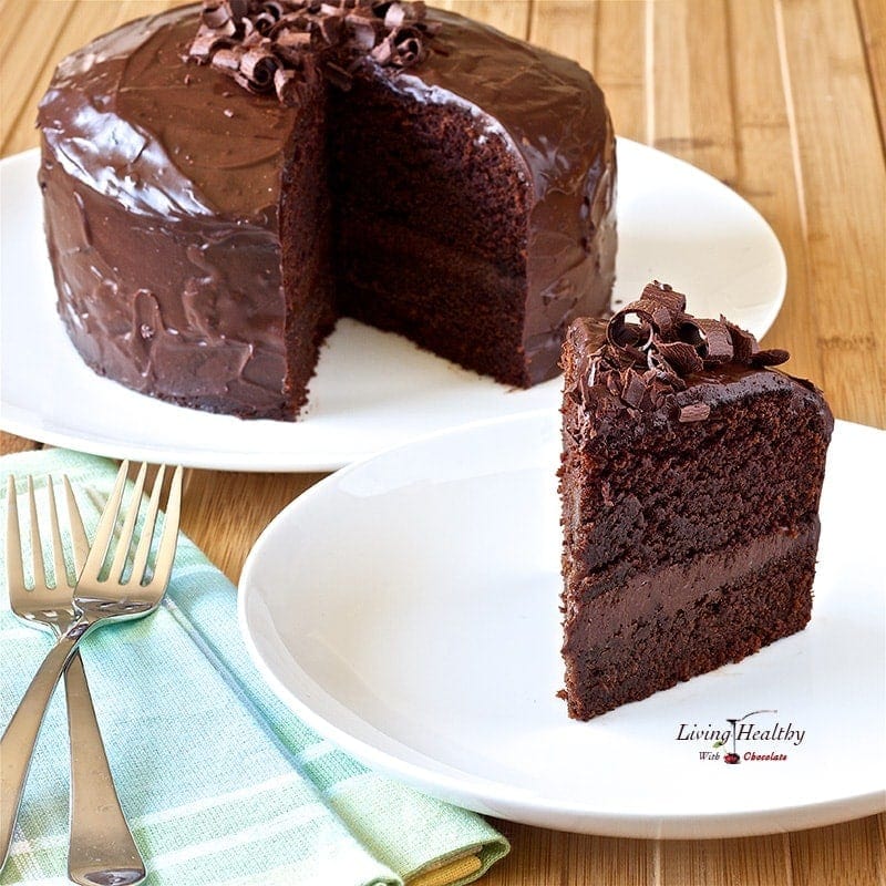 large slice of paleo chocolate cake in foreground with two forks on napkin and full cake in background 