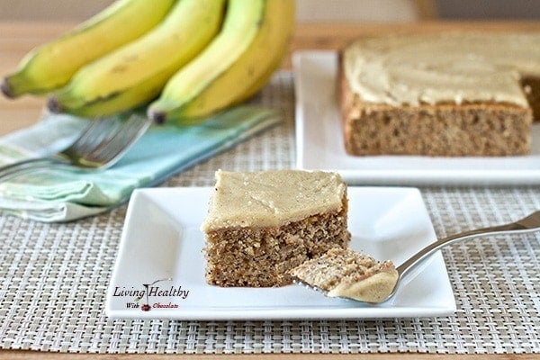 slice of banana poppy seed cake with white chocolate vanilla bean frosting on plate with more cake bananas in background