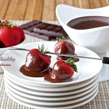 dark chocolate fondue covered strawberries on plate with bowl of melted fondue and loose strawberries in background