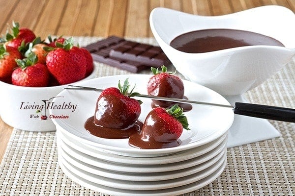 dark chocolate fondue covered strawberries on plate with bowl of melted fondue and loose strawberries in background 