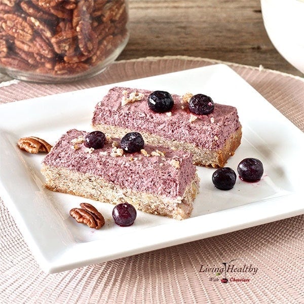 two blueberry breakfast bars on a white plate with loose berries on top and a jar of pecans in background