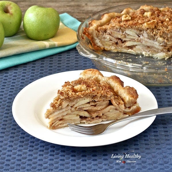 slice of homemade apple pie with a white plate and silver fork and glass dish behind with full pie and apples to the left