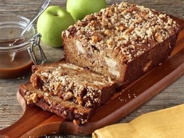 loaf of caramel apple pie bread on wooden cutting board with jar of caramel and two green apples in background