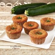 plate of five zucchini muffins on a brown and white place mat with two large zucchini in the background