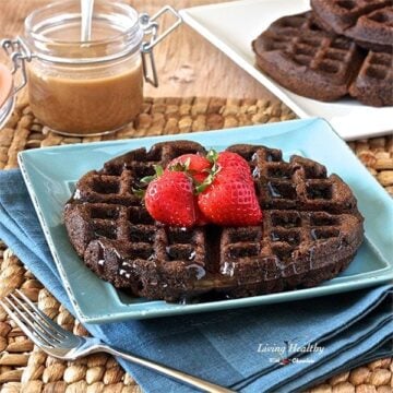 large chocolate waffle on a blue plate topped with fresh strawberries and nut butter and more waffles on plate in background