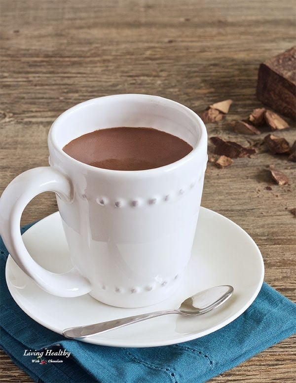 cup of hot chocolate served in white mug with small dish and blue napkin underneath and pieces of chocolate in background 
