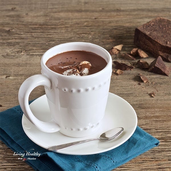 cup of hot chocolate served in white mug with small dish and blue napkin underneath and pieces of chocolate in background 