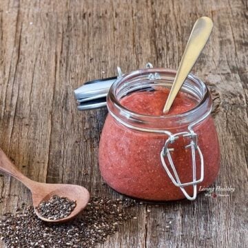 glass jar of chia seed strawberry jelly on a wooden table with spoon inside jelly and loose chia seeds and spoon next to jar