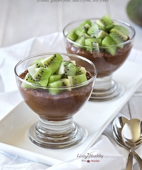 two glass serving bowls on a white plate filled with chocolate avocado pudding topped with chunks of kiwi