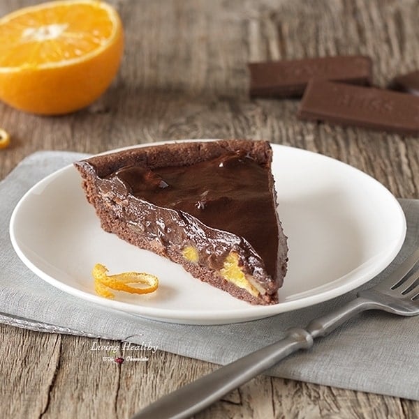 slice of dark chocolate orange almond tart on a white plate with a fork in front and half an orange and chocolate in back