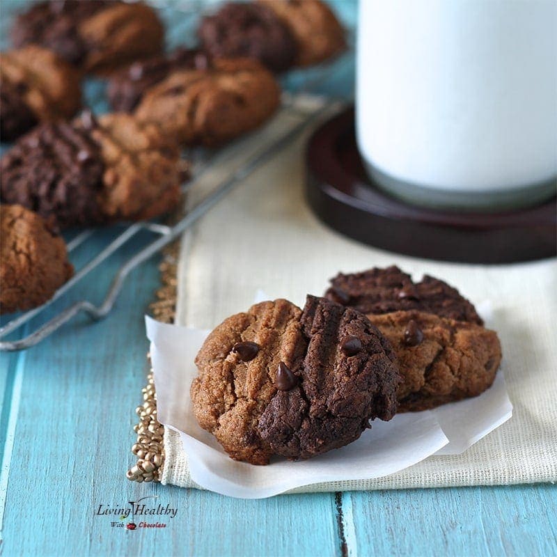 close up of two tones chewy chocolate cookies made with almond butter with a glass of milk and more cookies cooling in background