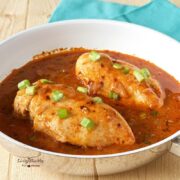 two chicken breasts laying in a bowl of spicy paprika sauce with scallions on top
