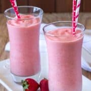 two glasses of strawberry milkshake with red straws served on white glass tray with a few loose strawberries.