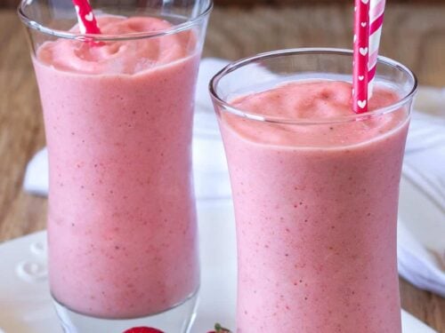 two glasses of strawberry milkshake with red straws served on white glass tray with a few loose strawberries.