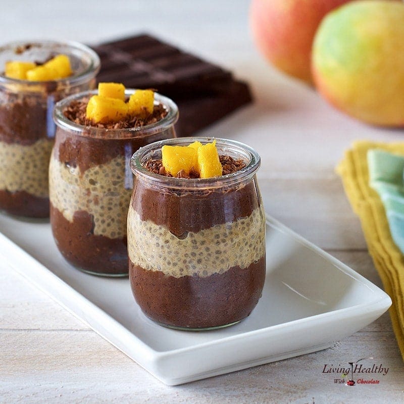 three serving jars with chocolate mango chia pudding on a white platter with mango and chocolate in background