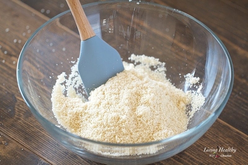 close up of a glass bowl of almond flour with spatula  sitting on wooden table