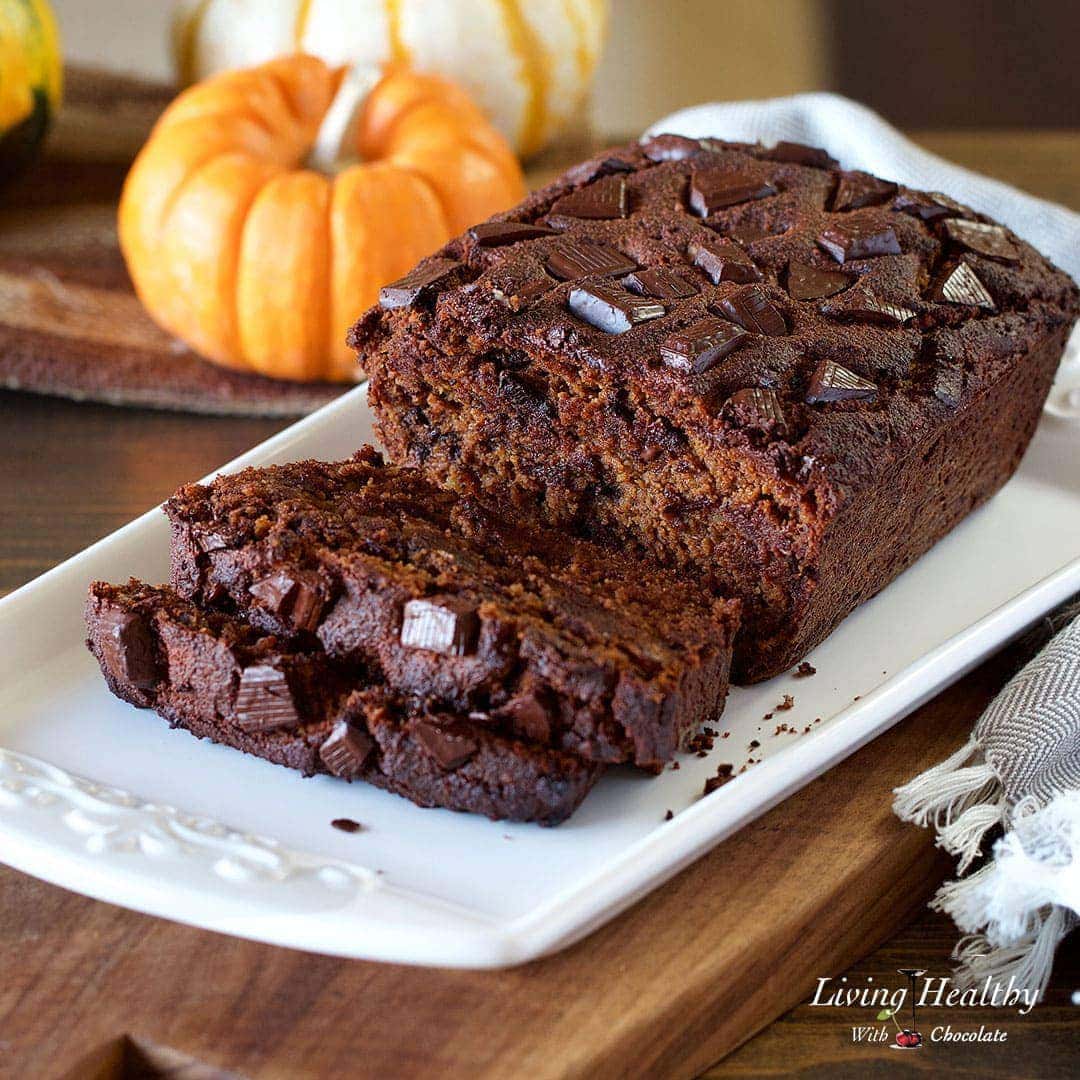 a loaf of chocolate pumpkin bread sliced and served on a white platter with small pumpkins in background