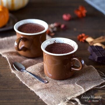 two cups of pumpkin hot chocolate sitting on a table in brown mugs with holiday decorations in the background