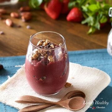 a glass filled with chocolate acai topped with homemade granola and two wooden spoons on a napkin next to the glass