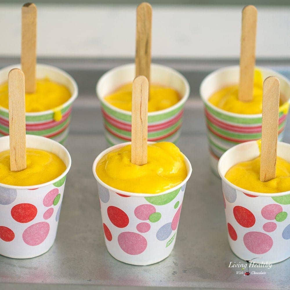 Six mango smoothie popsicles in dixie cups with popsicle sticks