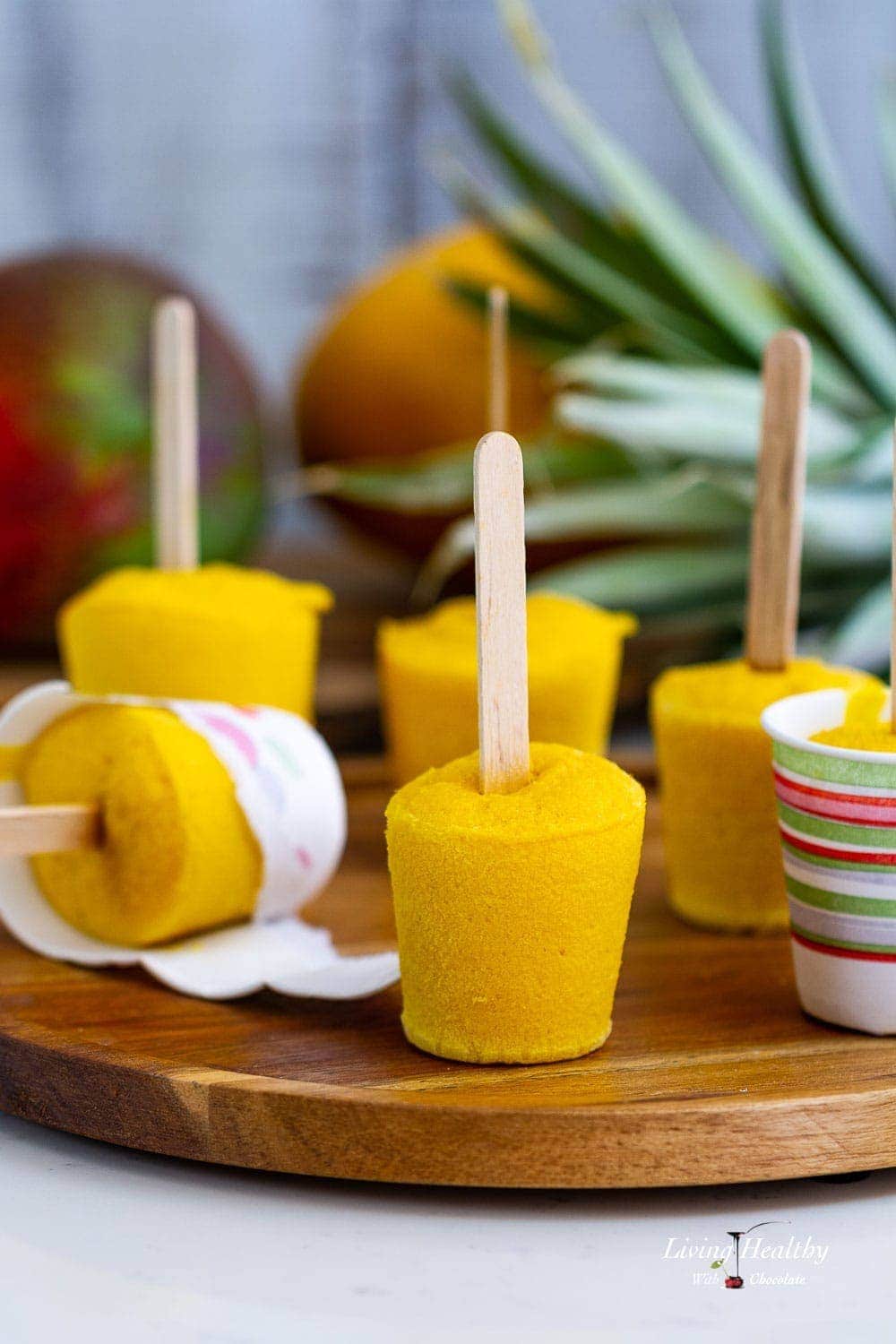 Mango smoothie popsicles on a plate with mango and pineapple behind them