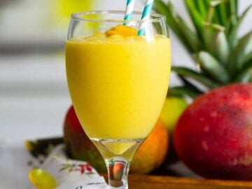 Glass of mango smoothie with two blue straws, plate of mango and pineapple behind the glass