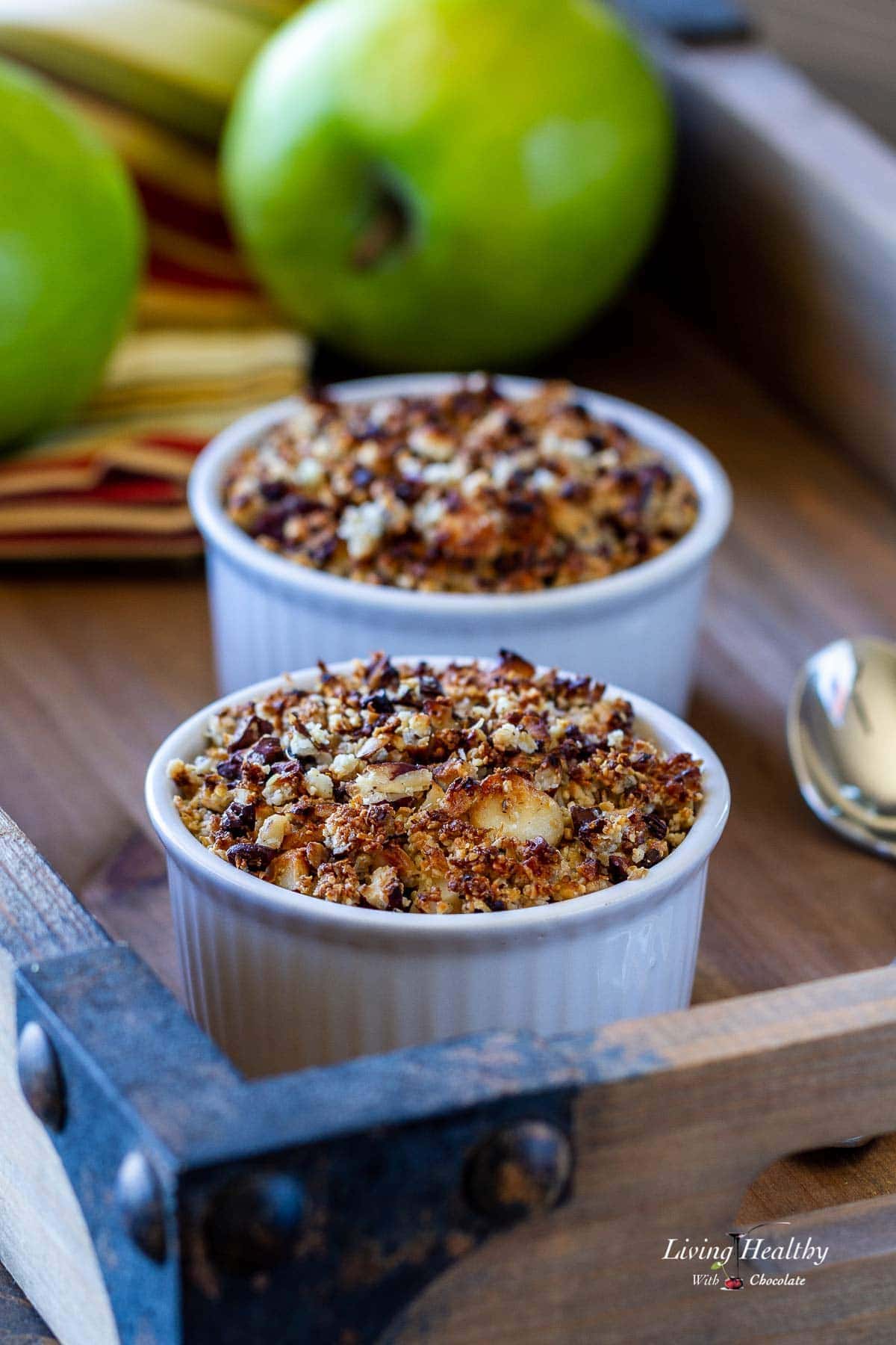 A tray with two ramekins of apple crisp and two green apples in the background sitting over a napkin 