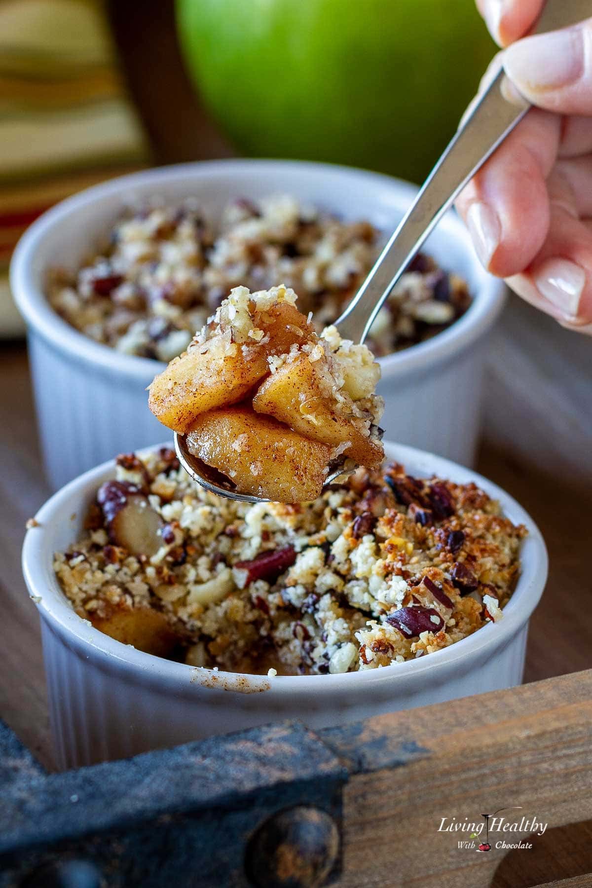 A spoon holding some apple crisp and two ramekins in a tray with an apple in the background