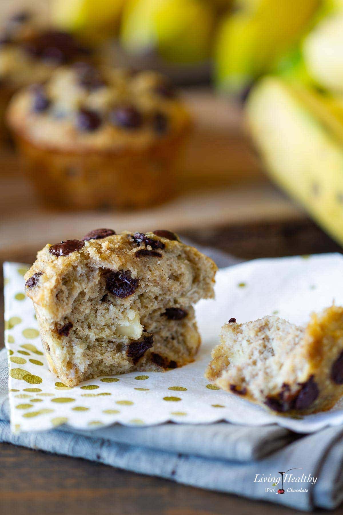close up of coconut banana muffin cut in half sitting on stack of multicolored napkins with banana and more muffins in background