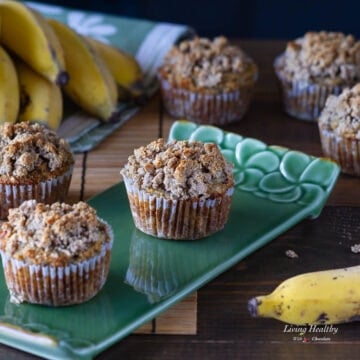 six banana muffins with crumb topping on a table and green plate around some yellow bananas