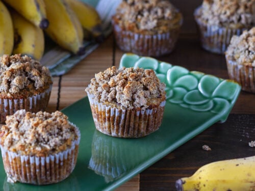 six banana muffins with crumb topping on a table and green plate around some yellow bananas