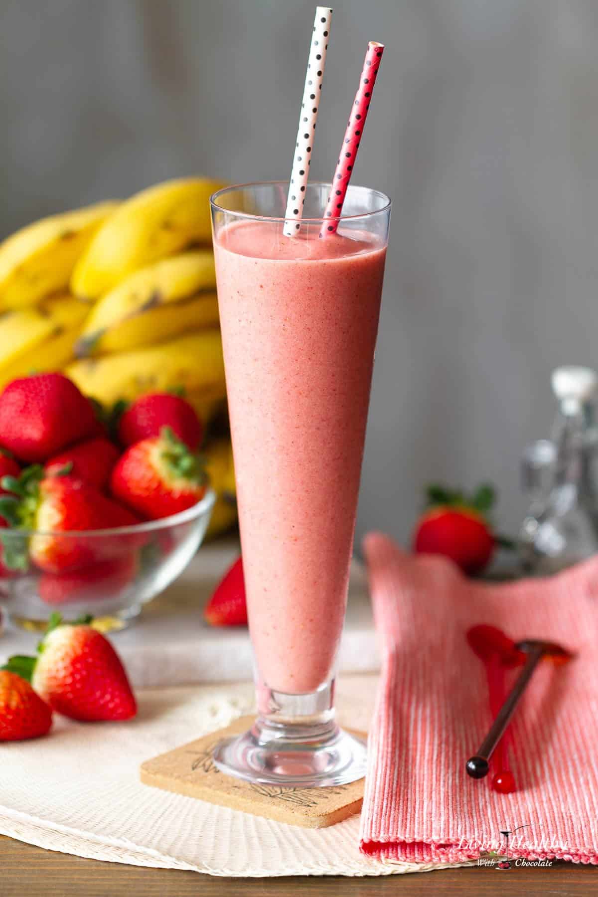 tall glass on a coaster filled with strawberry milkshake with two straws and some fresh fruits on the background.
