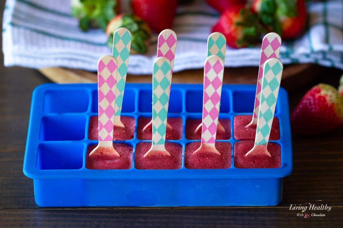 blue silicone ice cube tray filled with strawberry milkshake with a colorful popsicle stick in the center of each one.
