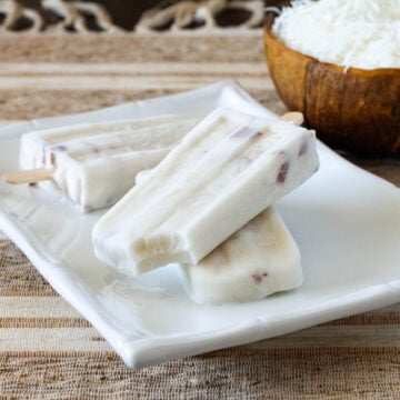 three coconut popsicles stacked on a square white plate with a coconut bowl behind it on a tan placemat
