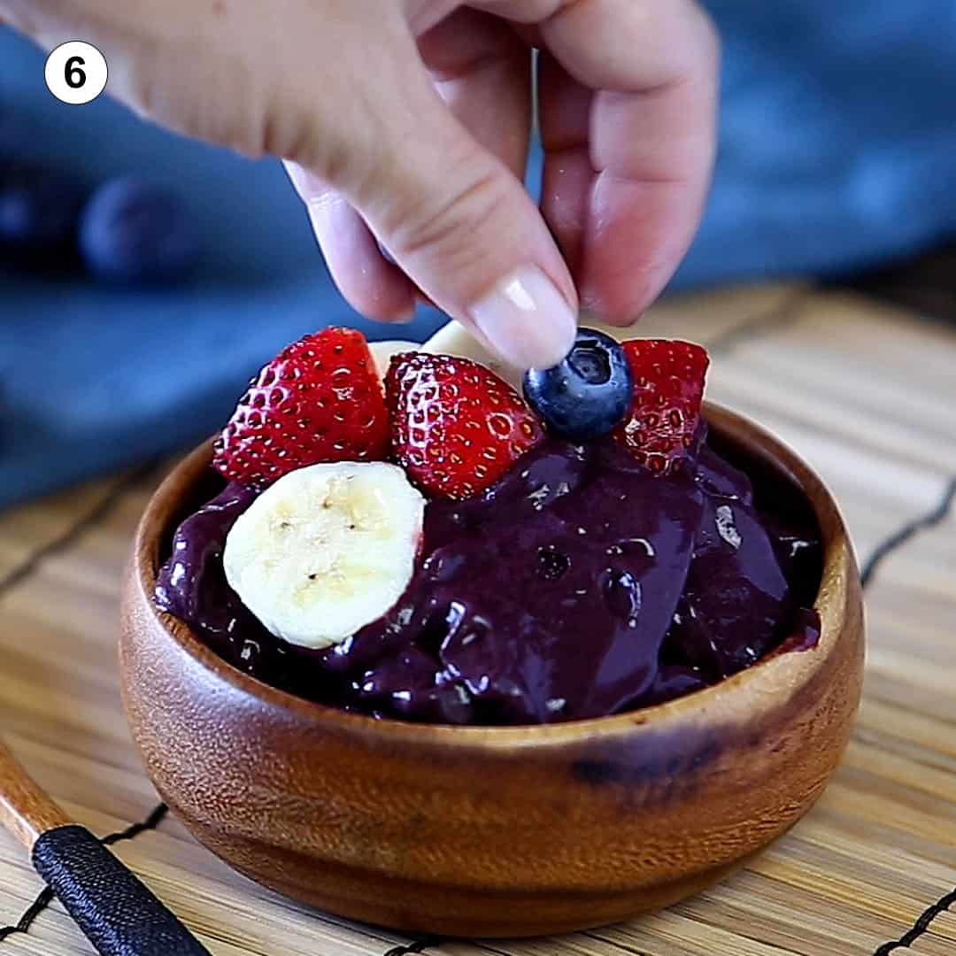 female hand placing a blueberry on top of an acai bowl topped with fresh sliced bananas and strawberries