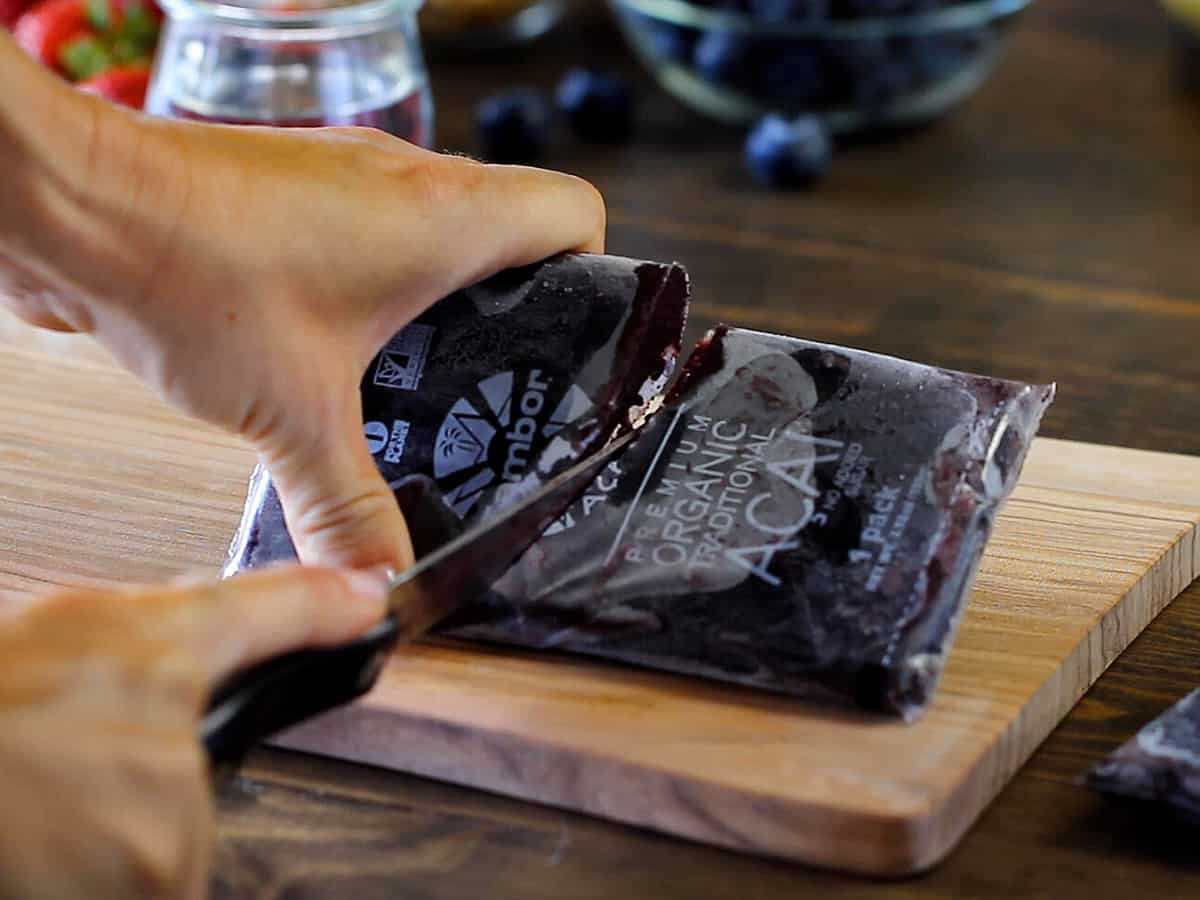 female hand cutting a pack of frozen acai puree in half with a knife on a wooden cutting board